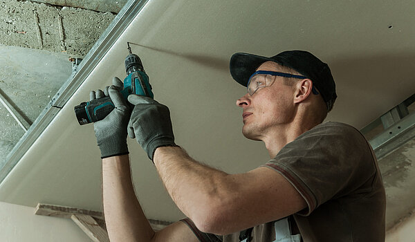 Mann mit Schutzbrille und Akkuschrauber befestigt ein Fertigelement an einer Dachwand